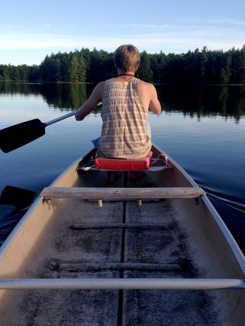 Person is seen from behind while canoeing on a tranquil lake with reflections of the surrounding forest visible on the water. The setting evokes a sense of peace, adventure, and connection with nature. This scene can be used for promoting outdoor activities, travel destinations, or wellness retreats.