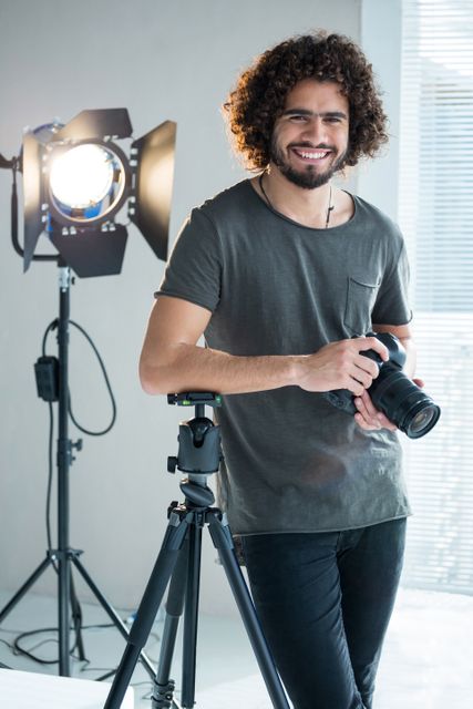 Young male photographer standing in a modern studio, holding a camera and smiling. Ideal for use in articles about professional photography, creative careers, or studio setups. Can also be used for promoting photography workshops, equipment, or lifestyle blogs.
