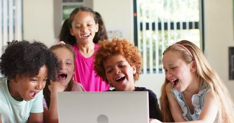 Laughing diverse children gathering around laptop, learning together in bright classroom