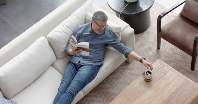 Man Relaxing on Sofa Reading and Reaching for Coffee Mug - Download Free Stock Images Pikwizard.com