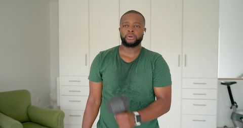 African man working out at home lifting dumbbell sweating wearing earbuds and smartwatch