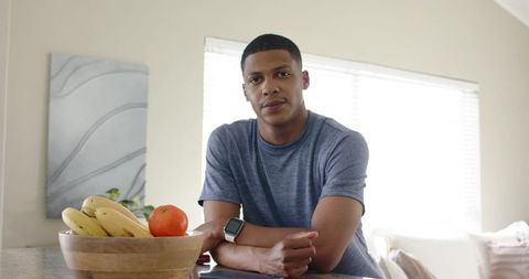 African american man leaning on kitchen counter, wearing smartwatch, with wooden bowl of bananas