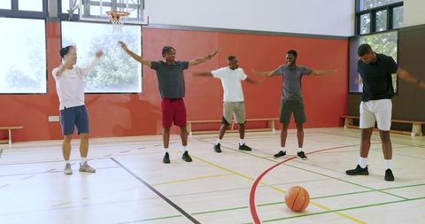 Diverse male basketball team warming up in gym following coach-led arm mobility drills