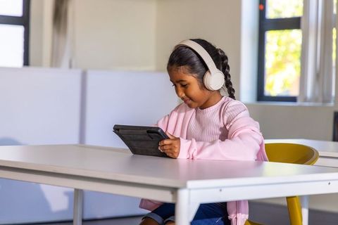 Asian schoolgirl wearing headphones using tablet at modern classroom desk for learning