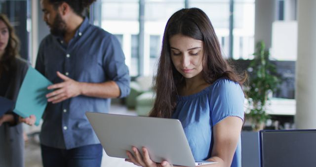 Young woman concentrating on laptop in modern office setting while coworkers discuss in the background. Useful for depicting modern business, teamwork, technology use in corporate environments, and professional women at work.