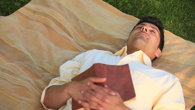 Man lying on grass-covered area using blanket for comfort, holding book Indicates leisurely outdoor activity, evokes sense of calm Can be used for themes related to relaxation, outdoors lifestyle, reading, disconnecting.