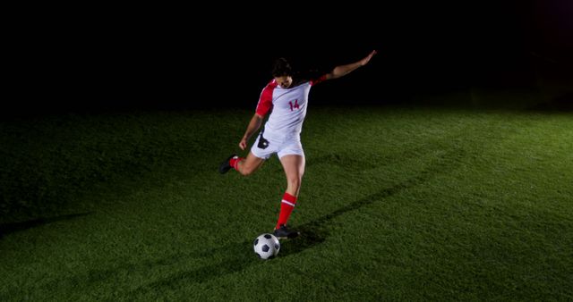 A young Caucasian male soccer player is kicking a ball on a dimly lit field at night, with copy space. His dynamic pose captures the energy and focus required in the sport, emphasizing the dedication to training and performance.
