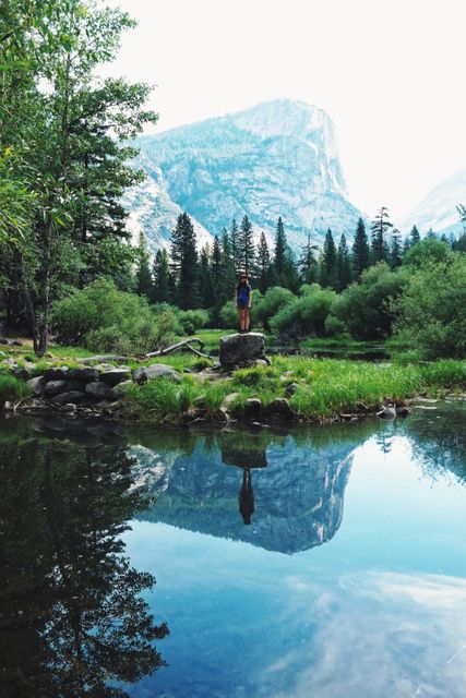 Person standing triumphantly on rock near calm lake reflecting nearby mountains. Ideal for outdoor adventure themes, promoting hiking destinations, nature conservation campaigns, travel blogs, and outdoor product advertisements.