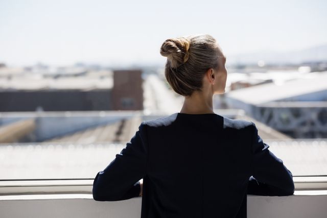 Businesswoman standing by office window, looking out thoughtfully. Ideal for themes related to professional life, career decisions, and business planning. Can be used for articles on work-life balance, leadership, and personal growth in the workplace.