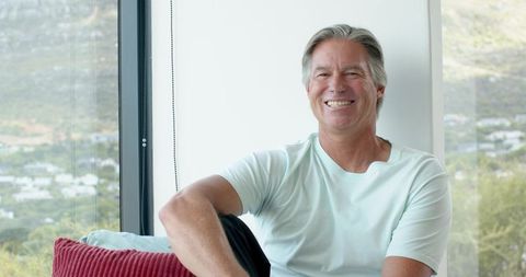 Relaxed senior man smiling while sitting by window with scenic view and cozy cushions