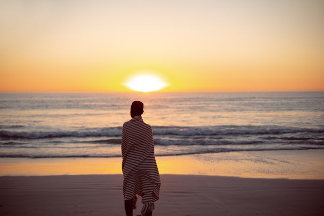 Rear view of woman wrapped in blanket standing on the beach during sunset