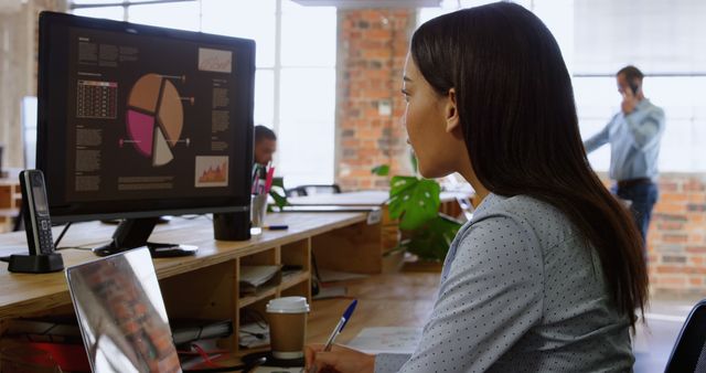 Businesswoman analyzing data on computer in modern office with brick walls and open space work environment. Ideal for use in articles, advertisements, or websites focused on business, technology, data analysis, office work, and professional settings.