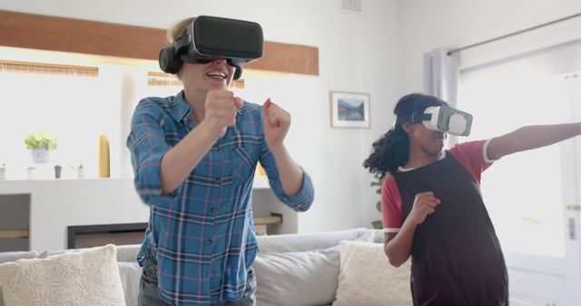 Two friends using VR headsets are enthusiastically playing a game in a living room. One is making a punching motion while the other is mid-jump, indicating excitement and engagement. Ideal for use in articles about VR technology, gaming communities, or modern entertainment at home.