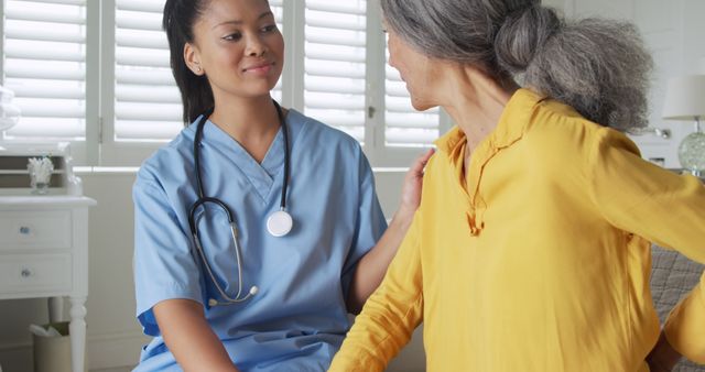Young biracial woman in nurse uniform at home, comforting elderly patient. She provides care and support in a comfortable home setting.