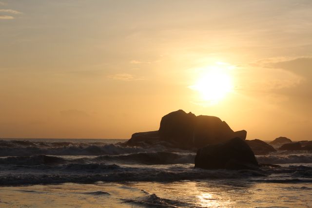 Silhouette of rocks during a golden sunset over an ocean beach. Waves lapping at the shore with soft, golden light creating a tranquil ambiance. Suitable for promoting relaxation, travel destinations, nature conservancy, or wellness retreats.