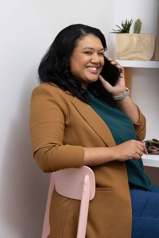Confident woman smiling talking on smartphone in minimalist home office, casual blazer