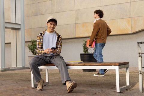 Casual college students sitting on campus bench using smartphone and carrying folders