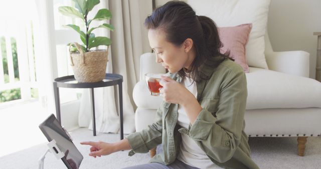 A woman is sitting on the floor in her living room, engaging with her tablet while holding a cup of tea. The setting is cozy with a comfortable white sofa, a pink cushion, a side table with a potted plant, and natural light shining in through the window. Ideal for lifestyle blogs, technology promotions, leisure time content, and modern home decor inspiration.