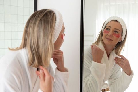 Woman in white robe applying under-eye gel patches while looking in mirror during skincare ritual