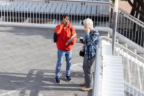Two young men talking on urban overpass holding notebooks and coffee in sunlight