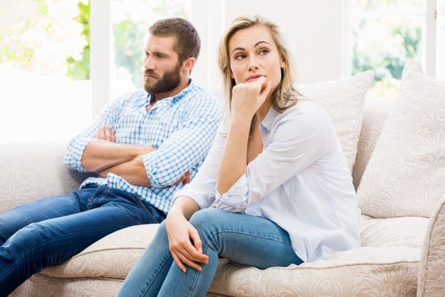 Young couple sitting on a couch in a living room, both looking away and ignoring each other. The scene suggests relationship conflict or tension. Useful for articles or content related to relationship issues, communication problems, or emotional struggles in partnerships.