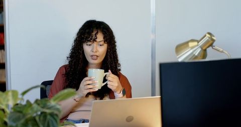 African american woman working from home office holding mug while using laptop and monitor