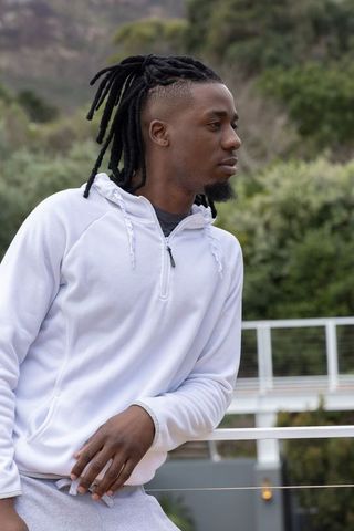 African american man leaning on terrace railing wearing white hoodie and gray joggers contemplative