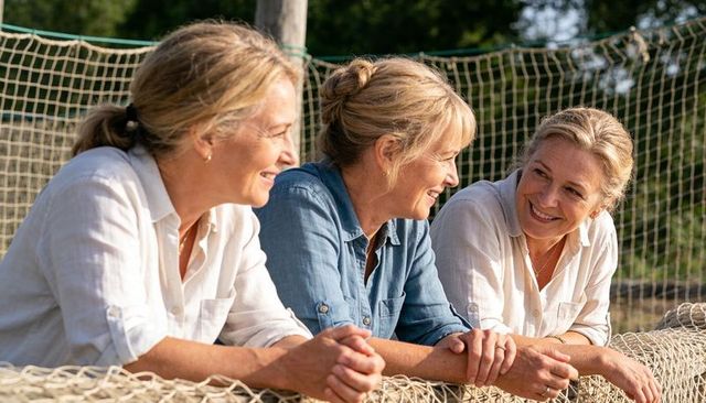 Three mature women leaning on net barrier smiling, chatting and laughing in sunlit park