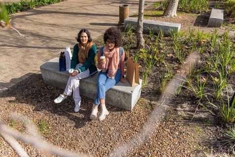 Urban friends sitting on concrete bench drinking coffee and chatting after shopping
