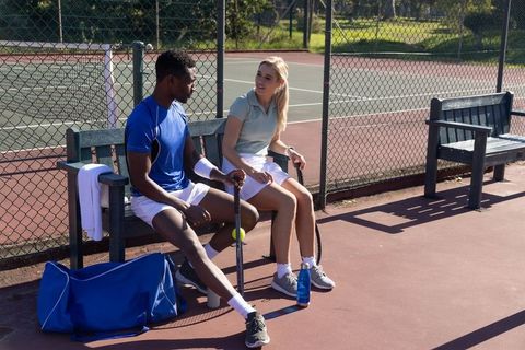 Diverse tennis players resting on bench and chatting after match at outdoor court
