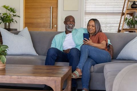 African american couple relaxing on sofa, browsing smartphone in cozy modern living room