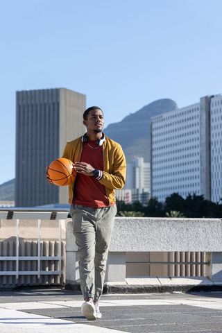 African american man walking on rooftop holding basketball with headphones, city skyline