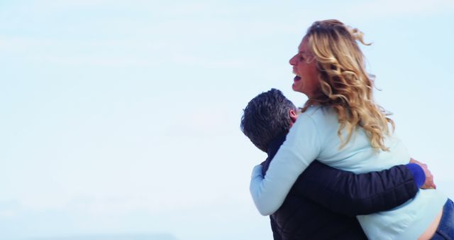A middle-aged Caucasian woman is embracing a man in a joyful hug against a clear sky backdrop, with copy space. Their cheerful interaction suggests a warm personal connection or a reunion.