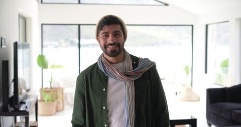 Smiling indian man standing in bright modern coastal living room wearing scarf and beanie
