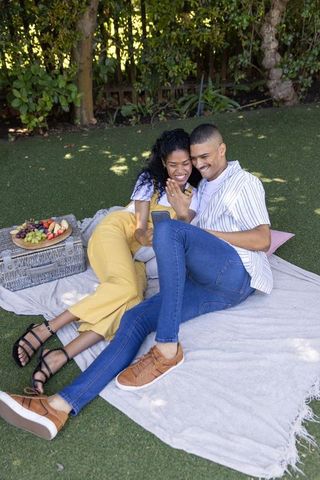 Couple laughing and sharing smartphone during relaxed backyard picnic