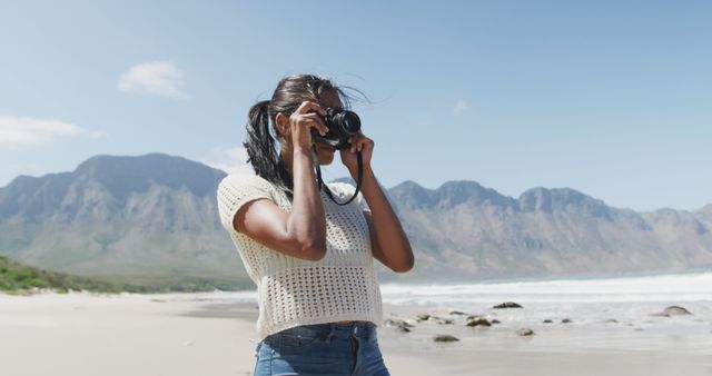 Young woman photographing beach scenery with mountains in background. Ideal for content on travel, nature explorations, hobbies, and leisure activities. Use in blogs, travel guides, tourism magazines, and photography tutorials.