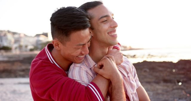Happy diverse gay male couple sitting and embracing by the sea at sunset. Summer, vacations, relationship, romance and free time, unaltered.