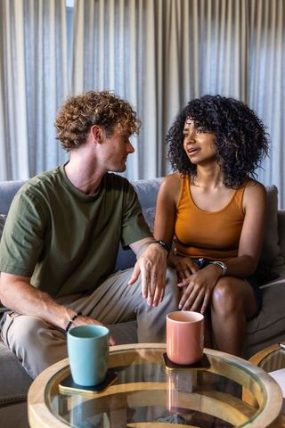 Diverse couple chatting on sofa with coffee mugs, sharing relaxed conversation in cozy living room