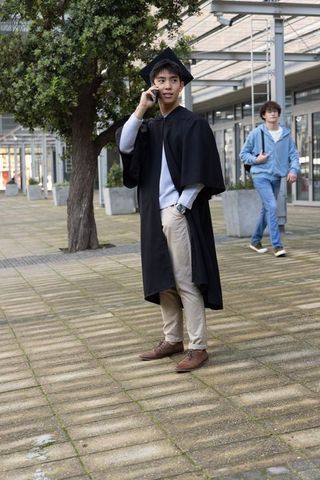 Young asian graduate talking on smartphone in modern campus courtyard wearing gown and mortarboard