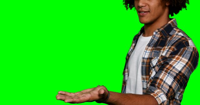Young man wearing a casual plaid shirt posing while holding out his open palm against a bright green screen background. The individual, not entirely visible except for the arm and partial torso, invites viewers to imagine what might appear on his hand. Useful for advertisements, digital presentations, or creative projects requiring green screen effects for the insertion of graphics or products.