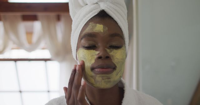 A woman is applying a facial mask while wearing a towel on her head in a spa-like bathroom. This is an ideal image for promoting skincare products, wellness routines, beauty blogs, and self-care guides. The serene and pampering vibe could also be used in advertisements for spa services or home spa products.