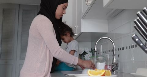 Mother in hijab washing dishes with daughter in kitchen