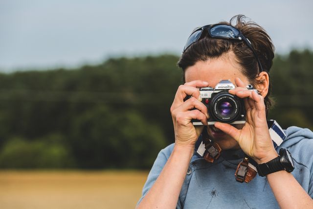 Person capturing nature scenes with film camera, standing outdoors in field, displaying passion for photography. Great for promotions on adventuring, hobbies, photography courses, travel blogs, and nature preservation.