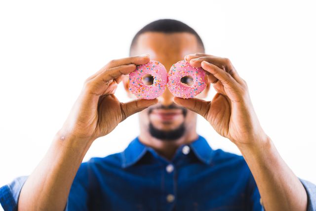This image shows an African American man holding two pink donuts with sprinkles in front of his face, creating a playful and fun atmosphere. The white background emphasizes the subject and the vibrant colors of the donuts. This image can be used for marketing campaigns related to desserts, sweet treats, or promoting a fun and joyful lifestyle. It is also suitable for social media posts, blog articles about indulgence, or advertisements for bakeries and confectioneries.