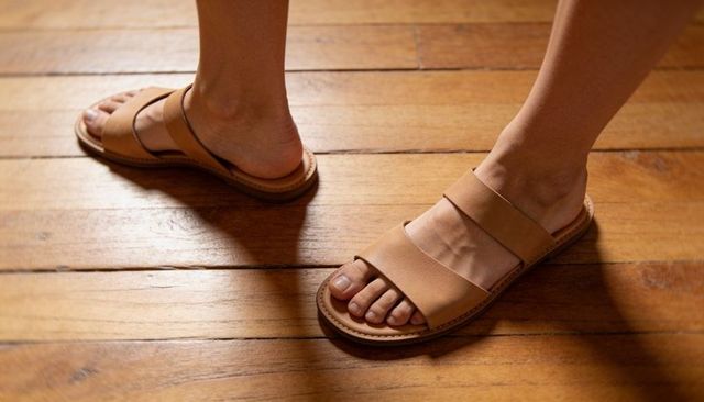Standing in tan leather slides on varnished hardwood floor with warm lighting and stitched sole