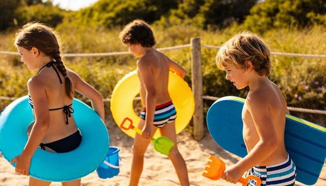 Kids walking to beach carrying colorful inflatables and boogieboard on sunlit sand dunes