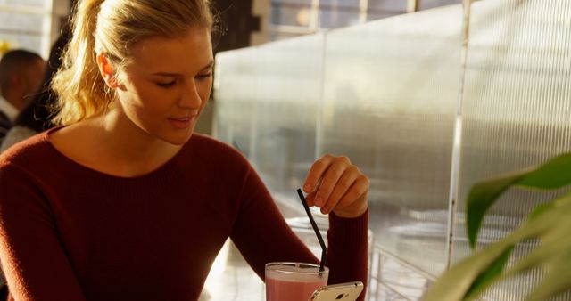 Young woman sitting at a cafe table enjoying a smoothie while looking at her mobile phone. She is wearing a maroon sweater and has her hair in a ponytail. The sun is shining through large windows, creating a warm and relaxed atmosphere. Ideal for illustrating concepts related to relaxation, casual dining, and modern technology use.