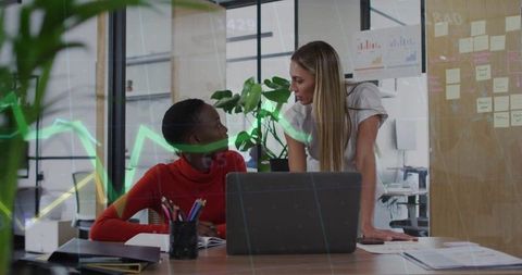 Two women collaborating and mentoring over laptop in modern open office with chart overlay