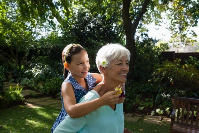 Senior woman carrying her granddaughter on her back in a lush, green backyard. Both are smiling and enjoying the moment, with the granddaughter holding flowers. Ideal for use in family-oriented advertisements, articles on intergenerational relationships, or promotions for outdoor activities and gardening.