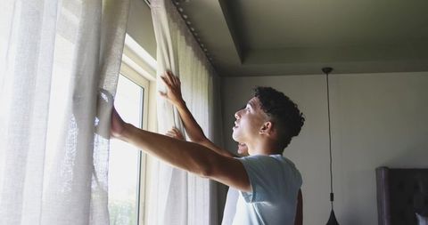 Young man opening curtains in sunlit bedroom morning routine natural light